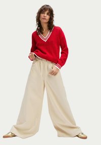 Roter gestrickter Pullover mit V-Ausschnitt und weißem Saum. Kombiniert mit lockeren kremfarbenen Hosen und Schuhen mit goldenen Akzenten.