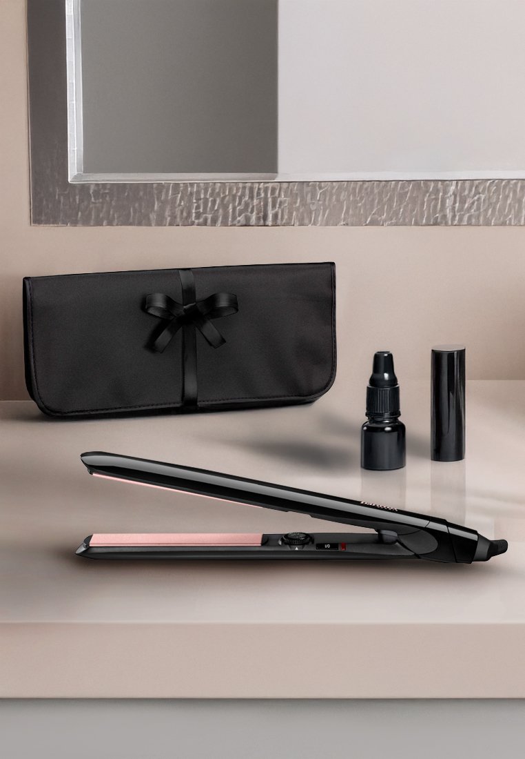 Fer à lisser en céramique avec un corps noir et des plaques roses, accompagné d'une housse de rangement noire, d'une bouteille de sérum et d'un spray de finition.