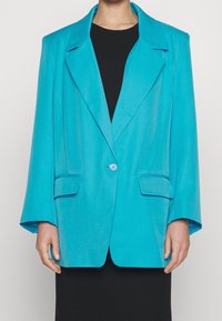 Turanžový blazer s jediným knoflíkem, širokými klopami a dvěma předními kapsami. Měkká textura a uvolněný střih, kombinovaný s černými šaty.