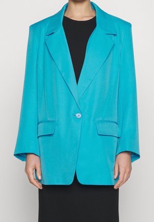 Blazer turchese con chiusura a un bottone, ampie alette e due tasche frontali. Tessuto morbido e vestibilità rilassata, abbinato a un vestito nero.