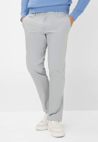 Man in een lichtgrijze rechte broek met handen in de zakken, gecombineerd met witte sneakers en een blauwe trui.