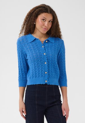 Femme portant un cardigan en maille texturée bleu vif avec des boutons en perle et des manches trois-quarts, associé à un jean taille haute foncé.