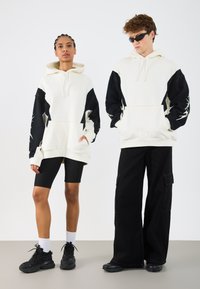 Capuchon truien in crème en zwart, met contrasterende mouwen en een crop ontwerp. Model combineert met zwarte shorts en sneakers.