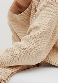 Béžový žebrovaný pletený svetr se širokým výstřihem a dlouhými rukávy, ukazuje texturovanou látku a volný střih. Vhodný pro každodenní nošení.