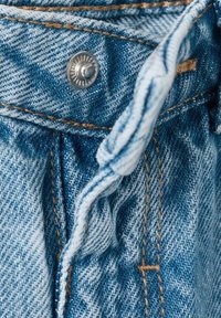 Jeans tkanina svetlo modre barve z vidnim kovinskim gumbom in kontrastnim rjavim šivom. Zloženi rob prikazuje teksturo in gosto tkanje.