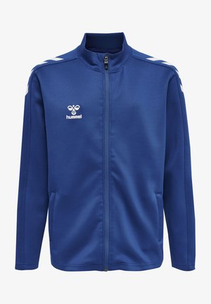 Blå atletisk jakke med høj krave, fuld lynlås, sidelommer og hvide chevron-striber på skuldrene. Har "Hummel" logo.