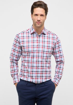 Mand iført langærmet skjorte med rødt, blåt og hvidt ternet mønster, kombineret med mørkeblå bukser, stående med hænderne i lommerne.