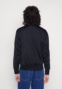 Svart satin bomberjacka med ribbat krage och cuffs, med en mjuk textur och enkel design. Bärs över blå denimbyxor.