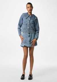 Denimu jaka ar lielu apkakli, priekšējām kabatām un pogu aizdari; kopā ar atbilstošu denimu svārku ar skalu malu. Melni apavi.