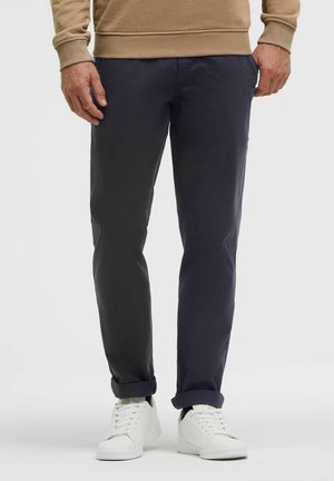 Mann trägt marineblaue Hose, weiße Sneakers und ein beiges Langarmshirt und steht mit entspannten Händen an den Seiten.