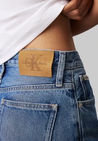 Vaqueros de mezclilla con un lavado en azul claro, que presentan un parche de cuero marrón con el logo "Calvin Klein Jeans" en la cintura y un diseño clásico de bolsillos.