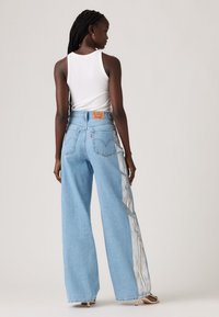 Woman with long braided hair wearing a white tank top and light blue wide-leg jeans with white side stripes, standing against a neutral background.