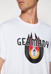 Hvid bomulds T-shirt med "TYSKLAND" i fed sort typografi og et cirkulært design med en fodbold og farvede accenter i rød, gul og sort.