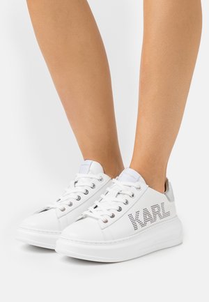 Białe skórzane sneakersy z płaską podeszwą, perforowanymi detalami oraz napisem "KARL" po boku, z metalicznym akcentem na pięcie.