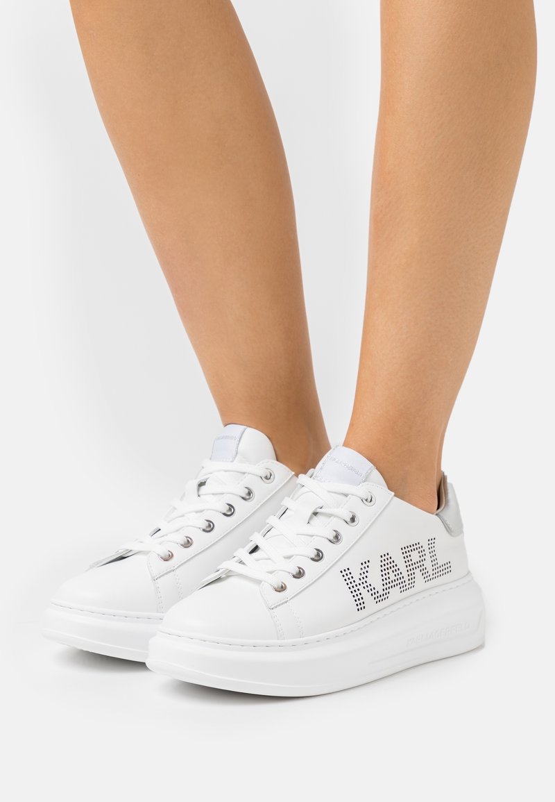 Białe skórzane sneakersy z płaską podeszwą, perforowanymi detalami oraz napisem "KARL" po boku, z metalicznym akcentem na pięcie.