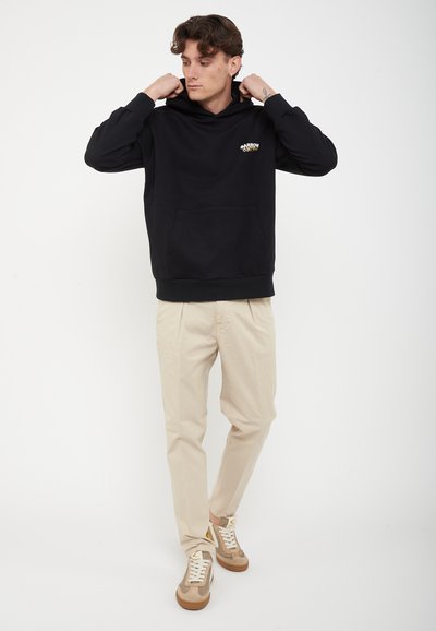 Νέος άνδρας που προσαρμόζει μαύρο φούτερ με μικρό λογότυπο, φοράει μπεζ παντελόνι και μπεζ αθλητικά παπούτσια, στέκεται μπροστά σε λευκό φόντο.