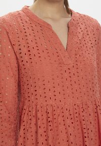 Korálové šaty s výstřihem do V, dlouhými rukávy a texturovanou látkou s otvorovou výšivkou. Sukně je stažena v pase.