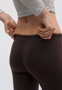 Pruunid elastsed kõrge vöökohaga retuusid, siledapinnaline tekstuur, keha järgiva lõike ja õmbluse detailidega. Modell kohandab vöökohta tagantpoolt.