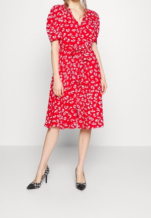 Woman wearing a red knee-length dress with white leaf patterns, puff sleeves, front buttons, and a waist tie, paired with black patterned heels.