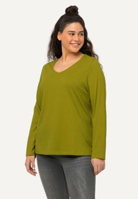 Frau mit dunklem, lockigem Haar, das hochgebunden ist, trägt ein olivgrünes Langarm-V-Ausschnitt-Shirt und graue Jeans, lächelt und schaut nach rechts.