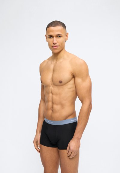 Μυώδης άνδρας με κοντά μαλλιά στέκεται μπροστά σε λευκό φόντο, φορώντας μαύρα μπόξερ με γκρι ζώνη μέσης και διακριτικό λογότυπο της μάρκας.