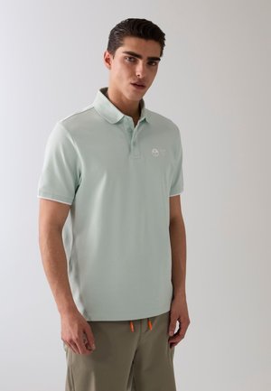 Jeune homme aux cheveux courts et foncés, portant un polo vert clair et un pantalon beige, debout devant un fond clair uni.