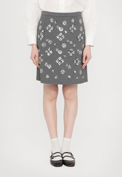 Jupe A-line grise avec des appliqués floraux argentés décoratifs. Portée avec des chaussettes blanches et des chaussures noires à double bride.