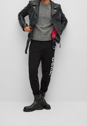 Person trägt schwarze Lederjacke offen, um das rote Futter zu zeigen, grauen Strickpullover, schwarze Hose mit dem Schriftzug "HUGO" und schwarze Stiefel vor einer einfarbigen Wand.