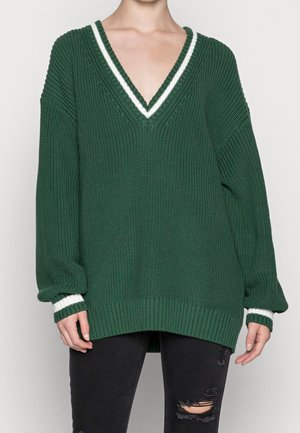 Personne portant un pull en maille vert foncé oversize avec des rayures blanches au niveau du col en V et des poignets, associé à un jean noir déchiré.