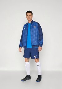 Blue athletic jacket with geometric pattern, blue t-shirt, blue shorts, white socks, and black soccer shoes. Nike logo on clothing.