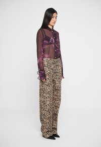 Prozirna ljubičasta košulja s cvjetnim uzorkom i volanima na rukavima, u kombinaciji s ležernim hlačama s leopard uzorkom u smeđoj i crnoj boji. Crne cipele s upitnim vrhom.