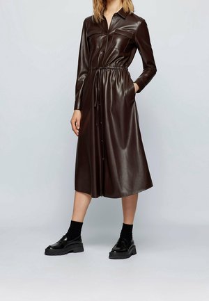 Bruine kunstlederen overhemdjurk met lange mouwen, elastische taille, voorzakken en knoopsluiting. Gecombineerd met zwarte loafers en sokken.