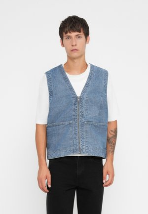 Denim vest in light blue, sleeveless with a deep V-neck, front zipper, two large pockets, and contrasting stitching. Worn over a white shirt.