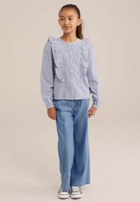 Mėlynas ir baltas dryžuotas marškinėliai su raukiniais priekyje ir ant rankovių, derinami su šviesiai mėlynomis plačiomis kelnėmis ir raštuotais sportbačiais.