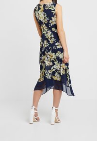 Marineblauwe bloemenjurk met een doorschijnende zoom, voorzien van gele en witte bloemmotieven, zonder mouwen en een rits aan de achterkant. Gepaard met witte bandjeshakken.