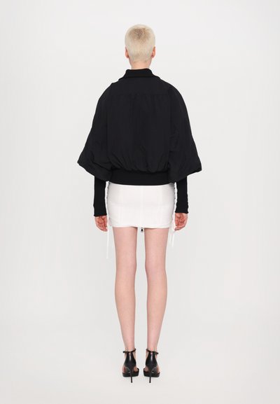 Jachetă neagră oversized cu mâneci bufante, tiv strâns, purtată peste o fustă mini albă. Modelul poartă sandale cu toc negre, întors cu spatele.