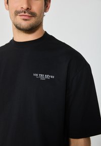 Black cotton t-shirt featuring a round neck and short sleeves, with white embroidered text on the left chest stating "VIS TES RÊVES LA TOUR EIFFEL PARIS."