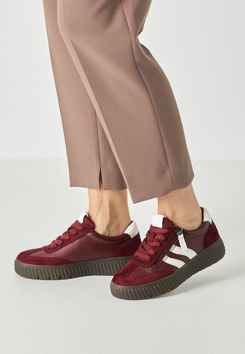 Pieds chaussés de baskets bordeaux avec des accents blancs et des semelles sombres, assortis à un pantalon beige court sur un fond uni.