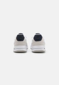 Hvite og lysegrå sneakers med en rund marineblå hældetalj, laget av semsket skinn og mesh-materialer, samt en teksturert gummisåle. Utviklet for komfort og stabilitet.