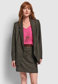 Mujer pelirroja con americana gris y falda abotonada con cinturón marrón, combinada con una camiseta gráfica rosa con un diseño de flamenco.