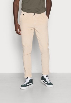 Pantalon en velours côtelé beige avec une coupe slim, poches avant et fermeture à bouton, associé à des baskets montantes vertes et un t-shirt vert olive foncé.