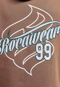 Tessuto marrone con audace testo ricamato bianco e teal "Rocawear" e il numero "99", con un design decorativo simile a fiamme.