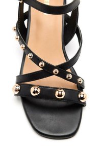 Sandalia de cuero negro con tiras entrecruzadas, que presenta acentos metálicos dorados y tachuelas redondas a lo largo de las tiras y la suela. Diseño de punta cuadrada.