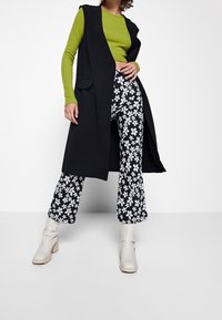 Lång svart väst över en grön långärmad topp, i kombination med vida byxor med svart och vitt blommönster och beige ankelstövlar.