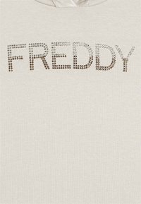 Sweatshirt beige chiaro con la scritta "FREDDY" in strass argento e oro lucidi sul petto. Texture liscia, design casual.
