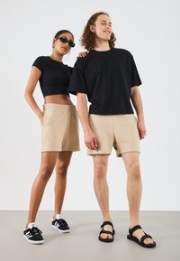 Crna skraćena majica i bež šorts za ženski model; crna oversized majica i bež šorts za muški model; oboje nose casual cipele.