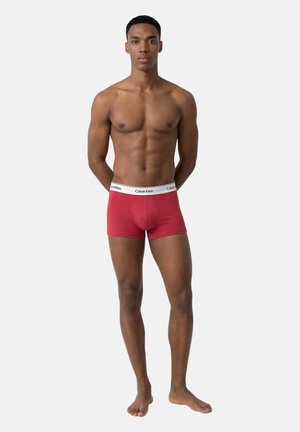Modelo masculino adulto de pie descalzo vistiendo calzoncillos bóxer rojos de Calvin Klein con cintura blanca con el logo de la marca sobre un fondo liso.