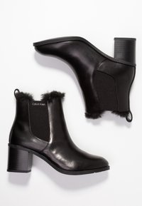 Botines negros de tacón con parte superior de cuero suave, paneles laterales elásticos, ribete de pelo sintético y tacón de bloque texturizado. Logotipo de la marca visible.