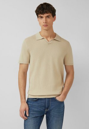 Jeune homme aux cheveux bouclés portant un polo à manches courtes beige texturé et un jean bleu, debout avec une main dans la poche.