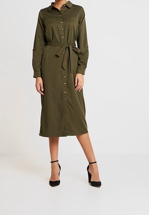 Kvinde iført en olivengrøn, langærmet, knap-ned midi-kjole med brystlommer, taljebånd og sorte ankelremssandaler.